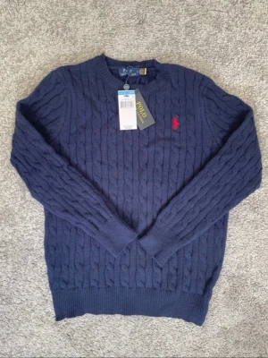Marinblå kabelstickad tröja Polo Ralph Lauren - Kabelstickad marinblå tröja från Polo Ralph Lauren med rund halsringning och röd broderad logotyp på bröstet. Tröjan har ribbade muddar vid ärmslut och nederkant. Klassisk och tidlös design i mjukt material.