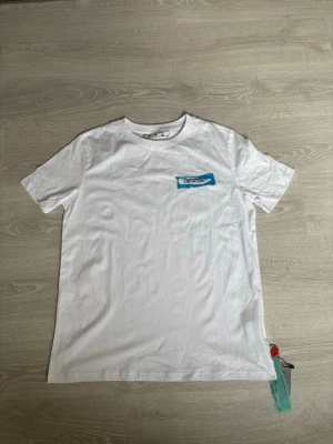 Vit Off-White t-shirt med blå logga - Säljer en vit t-shirt från Off-White med en blå logga tryckt på bröstet. Klassisk rund hals och korta ärmar. Tillverkad i mjuk bomull och har en avslappnad passform. Perfekt för dig som gillar streetwear och stilrena plagg. Stor i storlek!