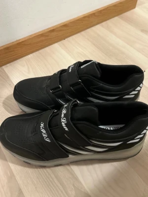Svarta sneakers med utfällbara hjul i 42 - Svarta sneakers från D-BuSun med två kardborreband och vita detaljer på sidorna. Skorna har en sportig look med perforerad tå och texten 'KDSP' på banden. Yttermaterialet ser ut att vara syntet och sulan är vit och svart.