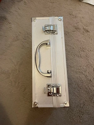 Silvrig aluminiumväska med handtag - Snygg och praktisk silvrig sminkväska i aluminium med metallhandtag och två lås framtill. Perfekt för att förvara smink, accessoarer eller andra småprylar. Väskan har en rektangulär form och stilren design som passar till det mesta.