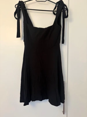 Svart kort klänning med knytband - Svart kort klänning med breda axelband som knyts upptill. Klänningen har en enkel, stilren design med lätt utställd kjol och fyrkantig ringning. Perfekt för dig som gillar minimalistisk stil och vill ha något bekvämt och snyggt.