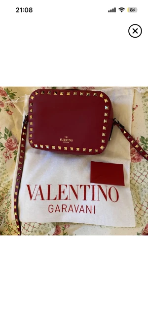 Röd axelväska Valentino Garavani - Säljer en snygg röd axelväska från Valentino Garavani med guldfärgade nitar runt kanterna. Väskan är i läder och har justerbar axelrem med nitar. Perfekt storlek för det viktigaste och ger en edgy vibe till din outfit.