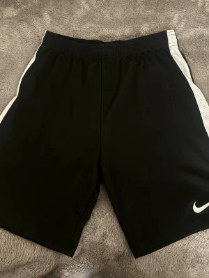 Svarta Nike shorts  - Svarta sportiga shorts från Nike med vita ränder längs sidorna och klassisk Nike-logga på ena benet. Perfekta för träning eller chill. Elastisk midja för skön passform och tillverkade i ett lätt och funktionellt material.