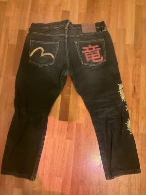 Svarta jeans med brodyr från Evisu - Unika svarta jeans från Evisu med broderade detaljer på bakfickorna, inklusive ett japanskt tecken och guldfärgad brodyr längs benet. Klassisk femficksmodell i denim med knappgylf och cool design som sticker ut. Limited edition 1/5 någonsin gjorda.