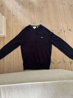 Mörkblå stickad tröja Lacoste - Mörkblå stickad tröja från Lacoste med V-ringning och den klassiska krokodilloggan på bröstet. Tröjan har långa ärmar och ribbade muddar vid ärmslut och nederkant. Perfekt för en clean och stilren look.
