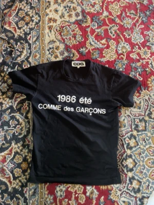 Svart Comme des Garçons t-shirt - Svart t-shirt från Comme des Garçons med vit text '1986 été' och logga på bröstet. Klassisk rund hals och korta ärmar. Perfekt för dig som gillar streetwear och vill ha något stilrent men ändå unikt.