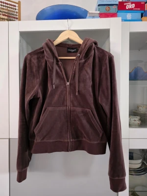 Brun velourhoodie från Juicy Couture - Mjuk och skön brun hoodie i velour från Juicy Couture. Dragkedja framtill, två stora fickor och justerbar huva med snören. Klassisk relaxed fit och ribbade muddar vid ärmar och nederkant. Perfekt för en chill och trendig look.
