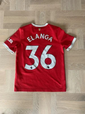Manchester United tröja Elanga - Röd Manchester United matchtröja från Adidas med Elanga och nummer 36 på ryggen. Tröjan har vita detaljer vid krage och ärmslut, samt klubbmärke och sponsorloggor på framsidan. Perfekt för fotbollsfans som vill stötta sitt lag. Fint skick, texten har släppt pyttelite på ryggen, skriv för mer info. Storlek 164 