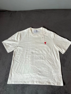 Vit t-shirt från Ami  - Stilren vit t-shirt från AMI Paris med det ikoniska röda hjärtat på bröstet. T-shirten är helt oanvänd och säljs med originaltaggar samt QR-kod för äkthetsverifiering kvar.  Skick  * Oanvänd med lappar * Två små hål/tråddragningar framtill på magen * Påverkar inte användningen och kan åtgärdas av den som vill  Detaljer  * Färg: Vit * Originaltaggar medföljer  Priset är satt med hänsyn till skadan, vilket gör detta till ett bra tillfälle att köpa en eftertraktad AMI Paris T-shirt!