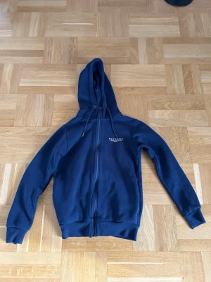 Marinblå hoodie från McKenzie - Snygg marinblå hoodie från McKenzie med dragkedja framtill och justerbar huva. Tröjan har ribbade muddar vid ärmslut och nederkant samt diskret logga på bröstet. Perfekt för en avslappnad och sportig stil. Har matchande byxor på profil