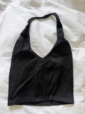 Svart ribbad halterneck linnetopp - Snygg svart ribbad linnetopp med halterneck och v-ringad urringning. Toppen är ärmlös och croppad, perfekt till höga jeans eller kjol. Materialet är mjukt och stretchigt, vilket gör den bekväm att bära hela dagen. Skickar samma dag.