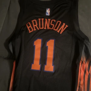 New York Knicks Brunson basketlinne - Snyggt svart basketlinne från Jordan med New York Knicks-tryck och Brunson 11 på både fram- och baksida. Linnet har orange och blå detaljer samt NBA-logga upptill. Perfekt för basketträning eller att bära som streetwear.