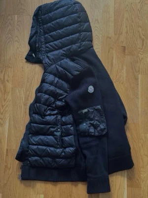 Svart pufferjacka från Moncler - Snygg svart pufferjacka från Moncler med stickade ärmar och huva. Storlek L men passar även M. Har blivit till användning 1-3 gånger inget fel.