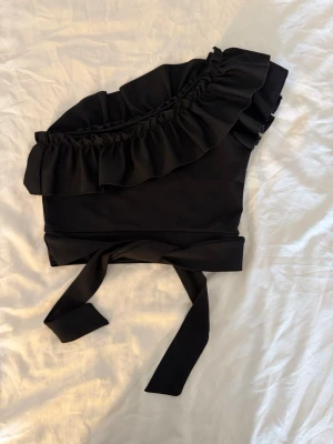 Svart oneshoulder topp med volang - Svart oneshoulder topp med bred volang längs halsringningen och knytband i midjan, går att knyta båda baktill och framtill. Passar xs och s