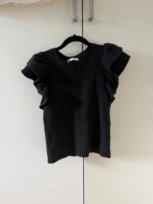 Svart ribbad topp med volangärmar - Snygg svart ribbad topp med volangdetaljer på ärmarna. Perfekt för dig som vill ha en enkel men ändå trendig look. Rund halsringning och korta ärmar med volang ger en extra touch. Passar till både jeans och kjol.