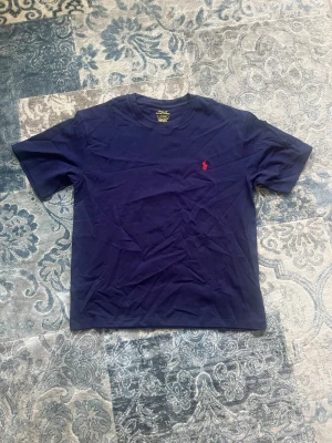 Marinblå t-shirt från Polo Ralph Lauren storlek S - Klassisk marinblå t-shirt från Polo Ralph Lauren med röd broderad logga på bröstet. T-shirten har rund halsringning och korta ärmar. Tillverkad i mjuk bomull för en skön känsla och enkel stil. Tröjan är storlek S. Den är helt ny och aldrig buren