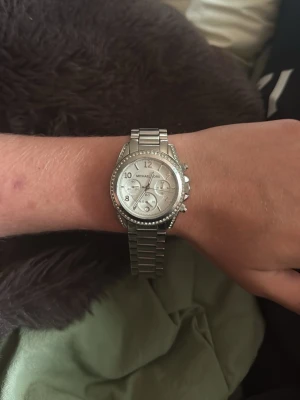 Silverfärgad klocka från Michael Kors - Stilren klocka från Michael Kors i silverfärg med länkarmband i metall. Urtavlan är rund och har flera mindre visare samt stenar runt boetten för extra bling. Perfekt accessoar för att lyfta din outfit. Nypris 2099kr 
