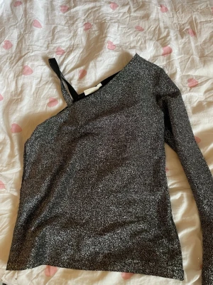 Glittrig oneshoulder topp i svart - Säljer en snygg svart glittrig topp med oneshoulder-design och lång ärm. Perfekt för dig som vill sticka ut med en cool och trendig look. Materialet är syntet och toppen har en slimmad passform som framhäver figuren.