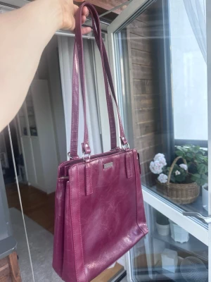 Vinröd axelväska i skinn - Snygg vinröd axelväska i äkta skinn med långa axelband och flera fack inuti. Väskan har en stilren, fyrkantig form och ljusbeige innerfoder. Funkar att andvända som ryggsäck också 