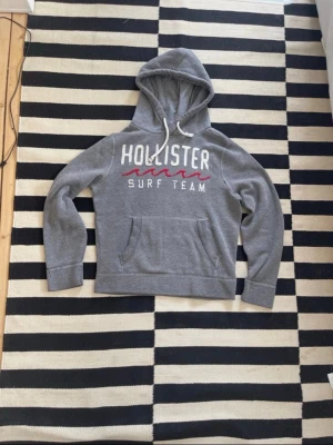 Grå Hollister hoodie med tryck - Snygg grå hoodie från Hollister med vit och röd text 'Hollister Surf Team' på bröstet. Klassisk känguruficka framtill, dragsko i huvan och mjukt material som känns skönt mot huden. Perfekt för chill dagar eller när du vill ha en avslappnad stil.