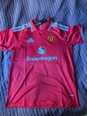 Manchester United matchtröja röd Adidas - Säljer en röd Manchester United matchtröja från Adidas med ljusblå detaljer och klubbmärke på bröstet. Tröjan har korta ärmar, klassisk krage och Snapdragons logga på framsidan. Perfekt för fotbollsfans som vill stötta sitt lag med stil.