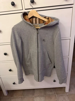 Grå zip hoodie från Burberry - Snygg grå hoodie från Burberry med dragkedja och klassiskt rutigt foder i huvan. Hoodien har fickor framtill och diskret Burberry-logga broderad på bröstet. Perfekt för dig som gillar stilrena och exklusiva plagg. Materialet är mjukt och bekvämt.snören vid luvan har gott av. 