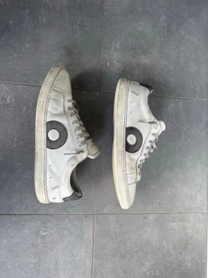 White low-top sneakers från Oliver Cabell - Snygga vita sneakers från Oliver Cabell med grå cirkeldetalj på sidan. Skorna har klassisk låg profil, vita snören och gummisula. Tillverkade med minimalistisk design och diskret logga på plösen.
