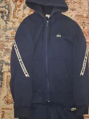 Mörkblå hoodie från Lacoste - Mörkblå hoodie med dragkedja från Lacoste. Klassisk krokodillogga på bröstet och vita band med Lacoste-tryck längs ärmarna. Huva med dragsko och två fickor framtill. Perfekt för en sportig och avslappnad stil. BYXOR INGÅR OCKSÅ👍