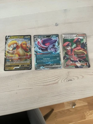 Pokémonkort Dragonite V, Gengar GX, Trainer - Tre Pokémonkort: Dragonite V med gul och orange design, Gengar GX med lila och blå färger samt ett Trainer-kort 'Pokémon Ranger' med röd och svart illustration. Samlarkort i klassisk Pokémon-stil, perfekta för dig som gillar TCG och anime.
