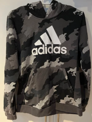 Adidas hoodie med grått camomönster - Snygg hoodie från Adidas med grått och svart camomönster och stor vit logga på bröstet. Tröjan har huva, magficka och ribbade muddar vid ärmslut och nederkant. Perfekt för en avslappnad streetstyle-look.