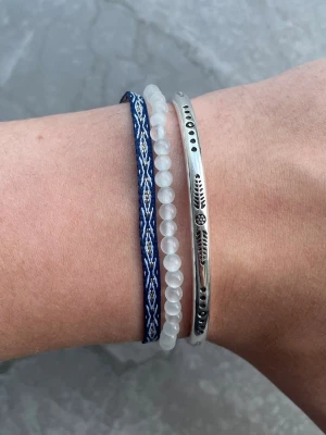 Armbands set - Säljer ett set med tre armband: ett i silverfärgad metall med ingraverade detaljer, ett med vita pärlor och ett blått band med vitt mönster. Perfekt för dig som gillar att mixa olika stilar och material. Alla armband är justerbara och sitter inte ihop men säljs som ett set. Perfekt till sommaren☀️
