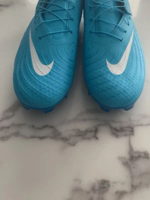 Nike Phantom GX blå fotbollsskor - Säljer ett par Nike Phantom GX fotbollsskor i en snygg ljusblå färg med vita detaljer och den klassiska Nike-loggan på sidan. Skorna har snörning och en modern, strömlinjeformad design som ger bra bollkontroll. Perfekta för dig som vill sticka ut på planen.