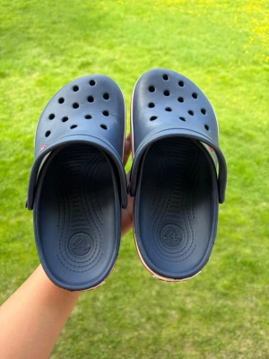 Mörkblå Crocs tofflor - Säljer ett par mörkblå Crocs tofflor med klassisk design och ventilationshål upptill. Tofflorna har en vit sula med röd rand och justerbar hälrem för extra komfort. Perfekta för chill dagar eller när du vill ha något enkelt att slänga på fötterna.