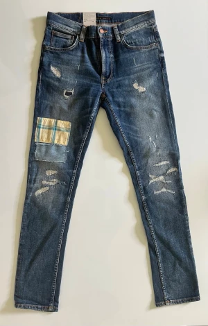 Blå slitna jeans med patch från Nudie - Säljer ett par blå jeans från Nudie med skinny fit och slitningar framtill. Unik patch med rutigt tyg och denim på vänster lår. Klassisk femficksmodell och orangea sömmar. Perfekta för dig som gillar en avslappnad och trendig stil.
