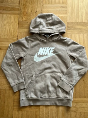 Beige hoodie från Nike med logga - Säljer en beige hoodie från Nike med stor vit logga på bröstet. Hoodien har huva, ribbade muddar och en praktisk magficka. Perfekt för dig som gillar sportig och avslappnad stil. Materialet är mjukt och bekvämt, idealiskt för chill dagar.