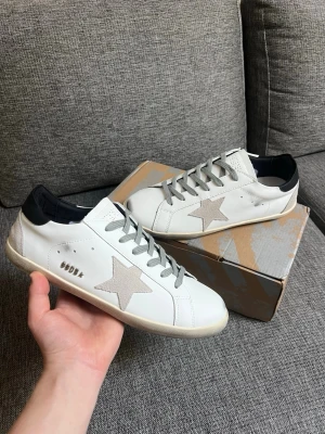 Golden Goose sneakers - Snygga vita sneakers från Golden Goose med grå mockastjärna på sidan, grå skosnören och svart hälparti. Skorna har en klassisk låg profil och är tillverkade i läder med detaljer i mocka. Perfekta för en trendig och avslappnad stil. Skorna är i storlek 43 och aldrig använda, säljer enbart pga de ej passade.