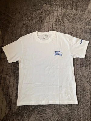 Vit Burberry t-shirt med blått tryck - Vit t-shirt från Burberry med blått tryck av riddare på bröstet och Burberry-logga på ärmen. Klassisk passform och rund halsringning. Tillverkad i mjuk bomull som känns skön mot huden. Perfekt för en clean och stilren look.
