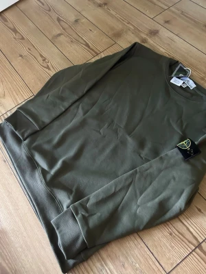 Olivgrön sweatshirt från Stone Island - Säljer en olivgrön sweatshirt från Stone Island med klassisk rund hals och ribbade muddar. Tröjan har den ikoniska Stone Island-loggan på vänster ärm och är tillverkad i mjukt bomullsmaterial. Perfekt för en avslappnad och stilren look.