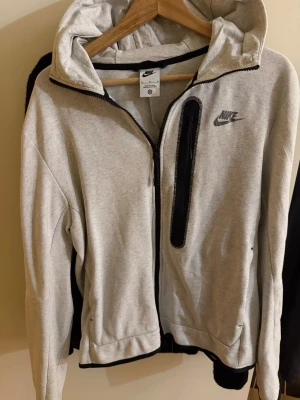Nike tröja - Snygg hoodie från Nike med svart logga på bröstet och dragkedja framtill. Hoodien har en huva och en svart detaljficka på bröstet. Tillverkad i mjukt sweatshirtmaterial och har ribbade muddar vid ärmslut och nederkant.