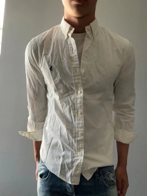 Vit skjorta från Ralph Lauren - Klassisk vit skjorta från Ralph Lauren med button-down krage och broderad logga på bröstet. Skjortan har lång ärm, knappar framtill och är tillverkad i bomull. Perfekt för en stilren och tidlös look.