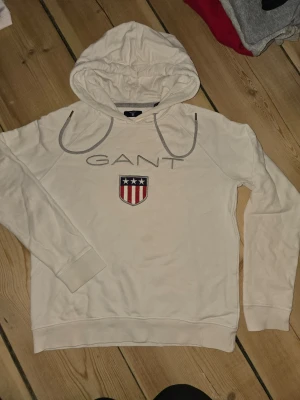 Vit hoodie från GANT med logga - Snygg vit hoodie från GANT med broderad logga och amerikanskinspirerat märke på bröstet. Hoodien har dragsko i huvan och ribbade muddar vid ärmslut och nederkant. Perfekt för en avslappnad och stilren look.