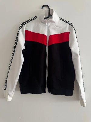 Vit, svart och röd hoodie med dragkedja - Snygg dragkedja i vitt, svart och rött med dragkedja framtill. Har vita ärmar med svartvitt mönster längs axlarna och en bred röd panel över bröstet. Två fickor framtill och hög krage. Perfekt för en sportig och trendig look.