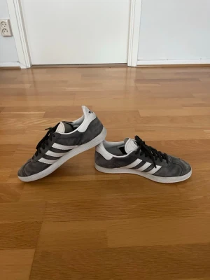 Adidas Gazelle sneakers – Storlek 41 - Säljer ett par stilrena och tidlösa Adidas Gazelle i grå mocka med vita detaljer. Skorna har den klassiska Gazelle-designen med tre vita ränder och vit gummisula som passar lika bra till vardags som till en mer uppklädd casual stil. Skorna är i fint begagnat skick med normalt slitage. Mockan har en snygg patina och sulorna har gott om liv kvar. Bekväma, lätta och enkla att matcha med de flesta outfits. Den exakta storleken är 40 2/3 men de är inprincip som storlek 41.