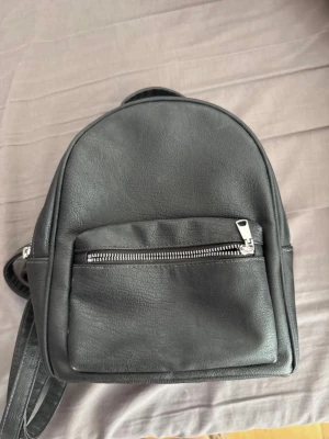 Svart minimalistisk ryggsäck - En stilren svart ryggsäck i fejkläder med justerbara axelband och en praktisk ytterficka med silverfärgad dragkedja. Perfekt storlek för det viktigaste och enkel att matcha med olika outfits.
