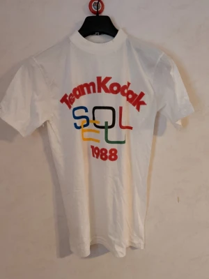 Team Kodak Seoul 1988 T-shirt - Vintage vit T-shirt med trycket 'Team Kodak Seoul 1988' i rött, blått, svart, gult och grönt. Klassisk passform med korta ärmar och rund hals. Perfekt för dig som gillar vintage och sportiga vibbar. Från. 1988, oanvänd, storlek Medium