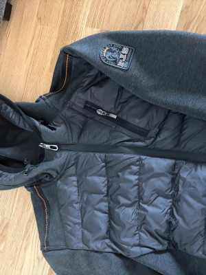 Mörkgrå pufferjacka från Parajumpers - Snygg mörkgrå pufferjacka från Parajumpers med vadderad front och mjuka ärmar i sweatshirtmaterial. Jackan har huva, bröstficka med dragkedja och en patch på axeln. Perfekt för kalla vinterdagar och riktigt stilren look.