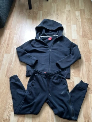 Nike tech - Svart loungewear set från Nike med zip-hoodie och matchande byxor. Hoodien har dragkedja, huva och diskret Nike-logga på bröstet. Byxorna har resår i midjan med dragsko och sidofickor. Perfekt för chill dagar hemma.