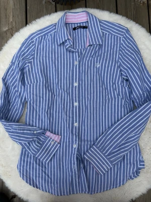 Blårandig skjorta från Gina Tricot (XS) - Snygg blårandig skjorta från Gina Tricot i bomull. Skjortan har vita knappar, lång ärm och en liten broderad logga på bröstet. Insidan av kragen och manschetterna har rosa och vita ränder som en extra detalj. Perfekt för en chill och stilren look.