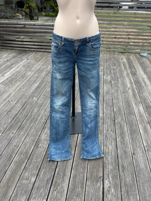 Blå bootcut jeans från LTB - Säljer ett par blå bootcut jeans från LTB med klassisk femficksdesign och snygga slitningar framtill. Jeansen har normal midja, raka ben och är gjorda i mjuk denim med kontrastsömmar. Perfekta för en avslappnad och trendig look.