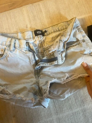Ljusa denimshorts från 157 - Säljer ett par ljusblå denimshorts från 157 med råa, fransiga kanter och slitna detaljer. Klassisk femficksmodell med bälteshällor och knappgylf. Perfekta för varma dagar och en avslappnad look.